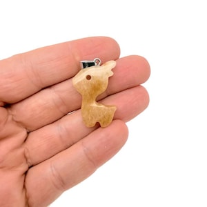 May include: A light brown pendant in the shape of a deer. The pendant has a silver-colored bail and a small hole near the top. The deer pendant is held in a hand against a white background.
