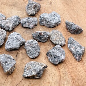 May include: A collection of 15 rough, gray and black, natural, tumbled,  crystals. The crystals are various sizes and shapes, and are arranged on a wooden surface.