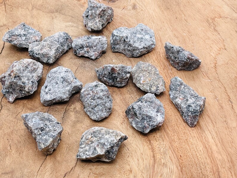 May include: A collection of 15 rough, gray and black, natural, tumbled,  crystals. The crystals are various sizes and shapes, and are arranged on a wooden surface.