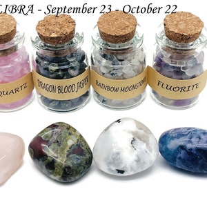 May include: Four small glass jars with cork tops filled with different colored crystals. The jars are labeled "Rose Quartz", "Dragon Blood Jasper", "Rainbow Moonstone", and "Fluorite".  Below the jars are four larger tumbled stones, one of each type of crystal.