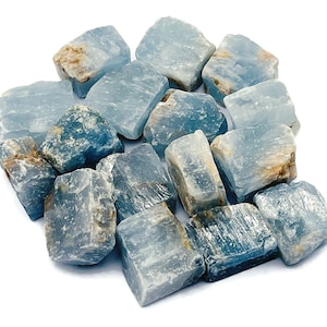 May include: A cluster of rough, pale blue calcite crystals. The crystals have a natural, unpolished look with varying shades of blue and some areas of tan. The stones are arranged on a white background.
