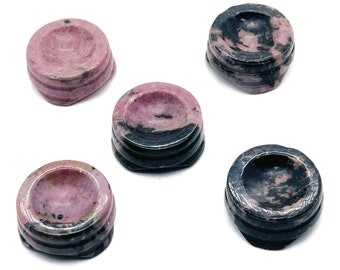 May include: Five small, round, pink and black, polished, natural stone coasters. The coasters are made of a layered stone with a smooth, polished surface.