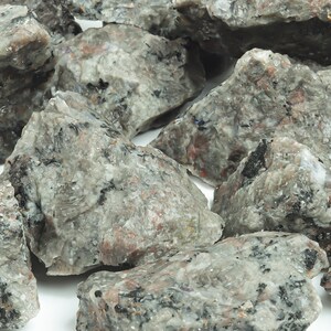 May include: A close-up of a pile of rough, gray, and black rocks with a speckled texture. The rocks are irregularly shaped and have a natural, unpolished look.