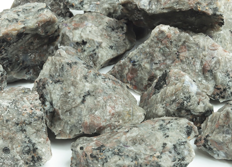 May include: A close-up of a pile of rough, gray, and black rocks with a speckled texture. The rocks are irregularly shaped and have a natural, unpolished look.