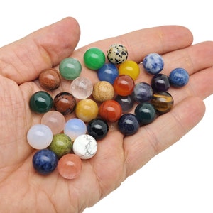 May include: A collection of 20 small, polished gemstone spheres in various colours, including green, blue, red, orange, yellow, brown, black, and white. The spheres are arranged in a hand, with some overlapping.