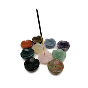 May include: A selection of flower-shaped incense holders in various colours, including red, green, and purple. An incense stick is inserted into a pink holder. The holders are made of polished stone.