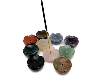 May include: A collection of flower-shaped incense holders in various colors, including red, green, and purple. An incense stick is inserted into a pink holder. The holders are made of polished stone.