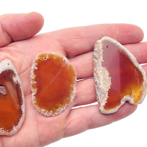 May include: Three agate slices with a hole drilled through the top. The agate is a light orange colour with white edges. The slices are on a hand.