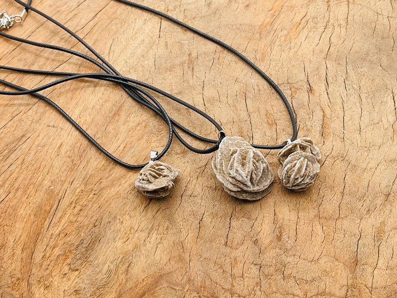 May include: Three rose quartz pendants on black cords. The pendants are different sizes and are arranged in a row on a wooden surface.