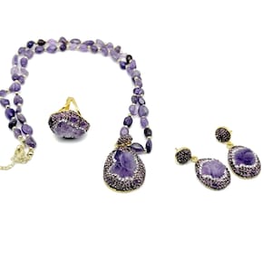 Può includere: Un set di gioielli in ametista viola con una collana, un anello e orecchini. La collana è realizzata con perline di ametista e ha un ciondolo con una pietra di ametista intagliata. L'anello e gli orecchini presentano anche pietre di ametista intagliate.