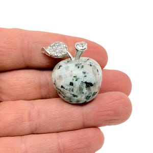 May include: A silver apple charm with a green leaf and a clear rhinestone on top. The apple is made of a speckled white and gray stone.