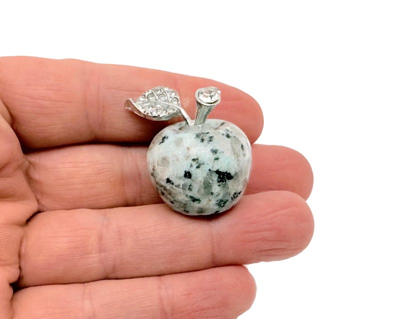 May include: A silver apple charm with a green leaf and a clear rhinestone on top. The apple is made of a speckled white and gray stone.