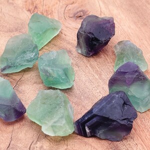 May include: A collection of green and purple fluorite crystals on a wooden surface. The crystals are rough and have a natural, unpolished look.