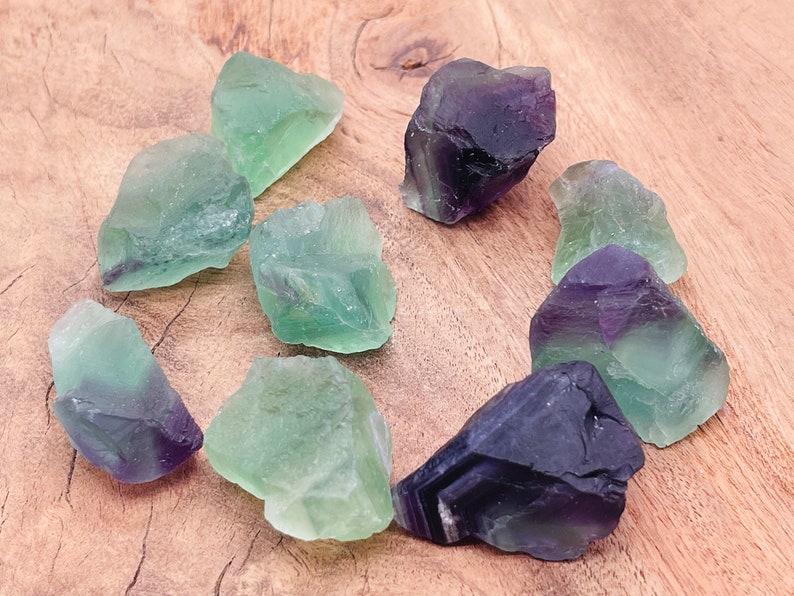 May include: A collection of green and purple fluorite crystals on a wooden surface. The crystals are rough and have a natural, unpolished look.