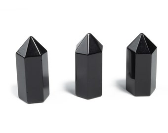 May include: Three polished black obsidian crystal points are arranged against a white background. Each crystal has a hexagonal base and a pointed top. The crystals are smooth and reflect light, showcasing their dark color.