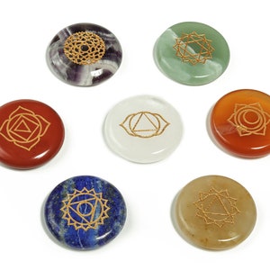 Könnte beinhalten: Sieben runde Chakra-Steine in verschiedenen Farben, darunter Rot, Orange und Blau. Jeder Stein zeigt ein goldfarbenes Chakra-Symbol. Die Steine sind auf weißem Hintergrund angeordnet, um ihre Farben und Designs hervorzuheben.