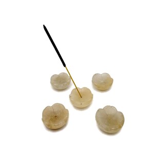 May include: Five flower-shaped incense holders in a light beige hue. One holds a black incense stick with a gold stem. The holders, made of a translucent material, are arranged on a white background, ideal for incense sticks.