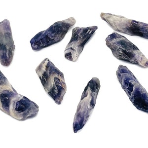Pode incluir: Uma coleção de oito pontas de cristal de ametista, cada uma com uma forma única e um tom roxo. Os cristais estão dispostos sobre um fundo branco.