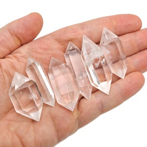 K&ouml;nnte beinhalten: Sechs klare Quarzkristallspitzen, auch bekannt als doppelt terminierte Quarzkristalle, sind in einer Reihe auf einer Hand angeordnet. Die Kristalle sind glatt und haben an beiden Enden eine spitze Spitze.