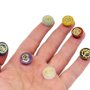 Może przedstawiać: Siedem kamieni czakr z złotymi symbolami na dłoni. Kamienie są niebieskie, zielone, żółte, pomarańczowe, czerwone, fioletowe i białe.