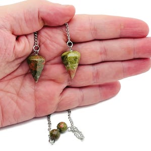 Op de afbeelding: Twee unakiet pendels met zilveren kettingen. De kegelvormige pendels vertonen groene en rood-oranje tinten. De kettingen zijn bevestigd aan de bovenkant van de pendels. Ze worden in een hand gehouden.