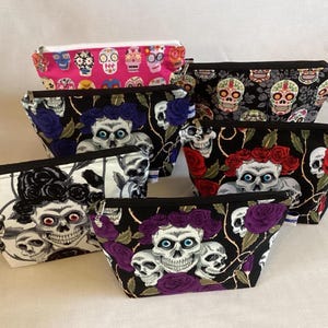 May include: A collection of zippered cosmetic bags with skull and floral patterns. The bags feature various designs, including skulls with floral crowns, roses, and a pink bag with colorful skull motifs. The bags are made of fabric and have black zippers.