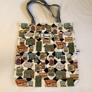 May include: A white tote bag with a black and white checkered pattern and a repeating pattern of vintage food tins and boxes. The bag has a black and white checkered handle and the word "Harrods" is printed on the front.