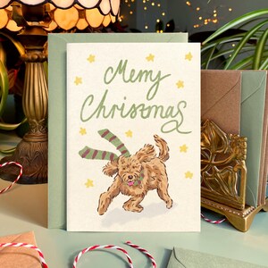 May include: A Christmas card with the words "Merry Christmas" in green script, featuring a playful brown dog wearing a green and red striped scarf. The card is surrounded by a green envelope and other decorative elements.