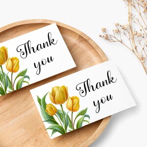 Puede incluir: Dos tarjetas blancas con el texto "Gracias" en letra cursiva negra. Una tarjeta presenta un ramo de tulipes amarillas con hojas verdes.