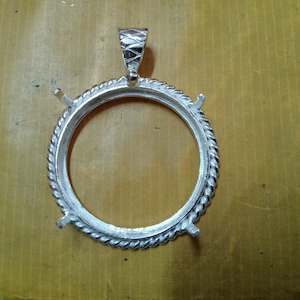 Könnte beinhalten: Silberner Anhänger-Fassung mit einer runden Zarge und vier Krappen. Die Zarge hat ein seilartiges Design am Rand.