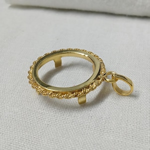 Puede incluir: Un colgante dorado con un marco circular y un borde texturizado en forma de cuerda. El colgante tiene tres pequeñas púas dentro del círculo y un lazo para una cadena. El colgante está sobre una superficie blanca.