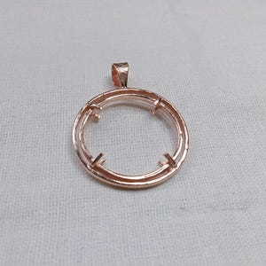 May include: A rose gold pendant setting with a circular design. The pendant has a loop at the top for a chain and four prongs to hold a stone. The setting is displayed on a white fabric background.