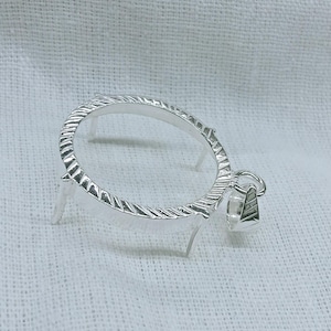May include: A silver-colored pendant with an oval frame and a textured design. The pendant has small legs and a small, geometric charm attached. The piece is displayed on a white, textured surface.