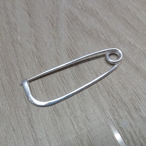 Può includere: Uno spillo da balia d'argento con un'ansa arrotondata nella parte superiore. Lo spillo è in metallo e ha una finitura liscia e lucida.