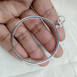 Könnte beinhalten: Ein silbernes Armreif-Armband mit einem runden, offenen Design und einem kleinen, dekorativen Anhänger an der Seite.