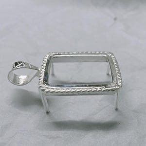 Puede incluir: Un colgante plateado con un marco rectangular y un borde decorativo en forma de cuerda. El colgante tiene un pequeño lazo para una cadena y cuatro patas pequeñas. Diseñado para sostener una piedra u otro inserto.