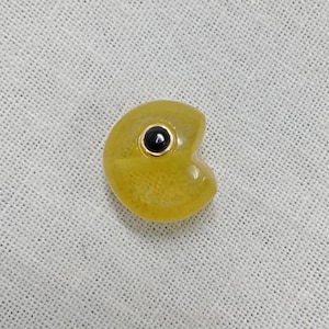 Pode incluir: Um pingente amarelo em forma de Pac-Man com uma pedra preta no centro.