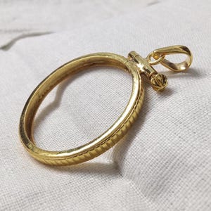 Puede incluir: Un colgante dorado con un diseño circular y un borde texturizado. Tiene un pequeño lazo para una cadena y un elemento decorativo en la parte superior. La joya se presenta sobre una superficie blanca.