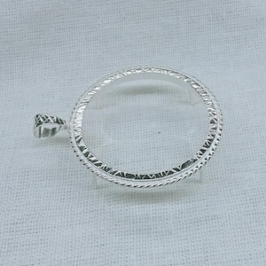 May include: A silver pendant frame, circular in shape. It has a textured, rope-style border and a decorative bail for a chain. The pendant is designed to hold a circular insert, such as a coin or charm.