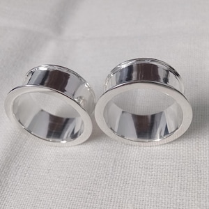 Può includere: Una coppia di tunnel per orecchie color argento lucido con una superficie liscia e riflettente e un design a bordo svasato. Questi gioielli circolari per il corpo presentano un centro cavo e una finitura metallica, dall'aspetto pulito.