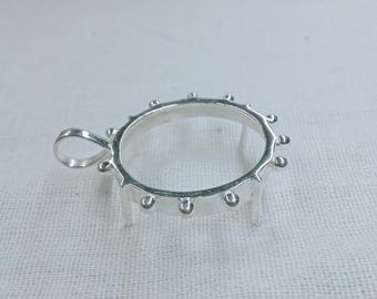 Könnte beinhalten: Ein silberner Anhänger mit kreisförmigem Design und kleinen dekorativen Kugeln am Rand. Der Anhänger hat eine Schlaufe für eine Kette und kleine Beine zur Unterstützung, vor einem weißen Hintergrund.