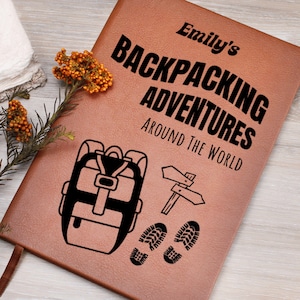May include: A brown leather journal with the text "Emily's Backpacking Adventures Around The World" in black lettering. The journal features a black silhouette of a backpack, a directional signpost, and footprints.