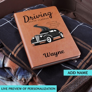 Peut inclure: Un carnet de planification d'itinéraire de conduite en cuir marron avec un dessin de voiture noire et le nom "Wayne" imprimé. Le carnet est posé sur une chemise à carreaux. Le texte "Ajouter un nom" et "Aperçu en direct de la personnalisation" sont également visibles.