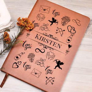 May include: A rose-gold leather journal with the name "KIRSTEN" printed in black. The journal is decorated with black line art of hearts, cupids, bows, and the words "Love you". A brown ribbon bookmark is visible.