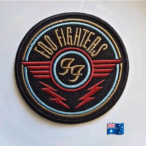 Puede incluir: Un parche circular negro con la inscripción "FOO FIGHTERS" en dorado. El diseño central presenta "FF" en dorado, alas rojas, detalles azules y rojos y dos rayos rojos. El parche es bordado.