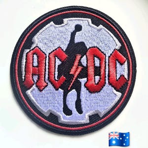 Puede incluir: Un parche bordado circular que presenta el texto rojo AC/DC con un rayo central. El diseño incluye una silueta negra de una persona sobre un fondo de color violeta claro en forma de engranaje, todo dentro de un borde negro grueso con ribete rojo.