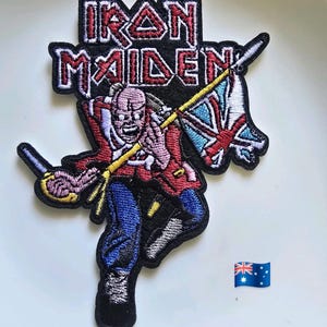 May include: An embroidered patch featuring the Iron Maiden logo in red and white text above a character in a red coat and blue trousers holding a flag with a Union Jack design. The patch has a black border and is shown next to an Australian flag icon.