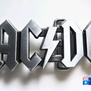 May include: A silver-toned metal belt buckle featuring the letters A, C, D, and C in a bold, angular font. A lightning bolt symbol is positioned between the letters C and D. The buckle has a brushed metallic finish and a rectangular loop attachment.