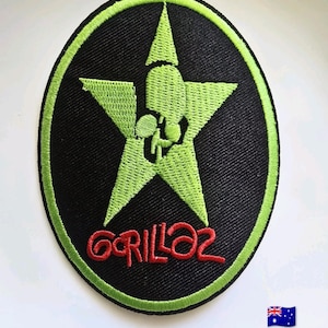 May include: An oval-shaped embroidered patch featuring a black background with a bright lime green border. In the centre, a lime green star contains a stylised silhouette graphic. Below the star, the word Gorillaz is stitched in red thread.