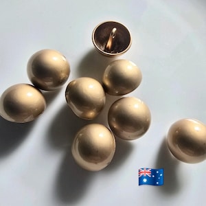 Op de afbeelding: Een verzameling van acht goudkleurige, ronde knopen. Zeven knopen zijn massieve bollen, terwijl één een lus aan de achterkant heeft. De knopen zijn gerangschikt op een wit oppervlak. De Australische vlag staat rechtsonder.